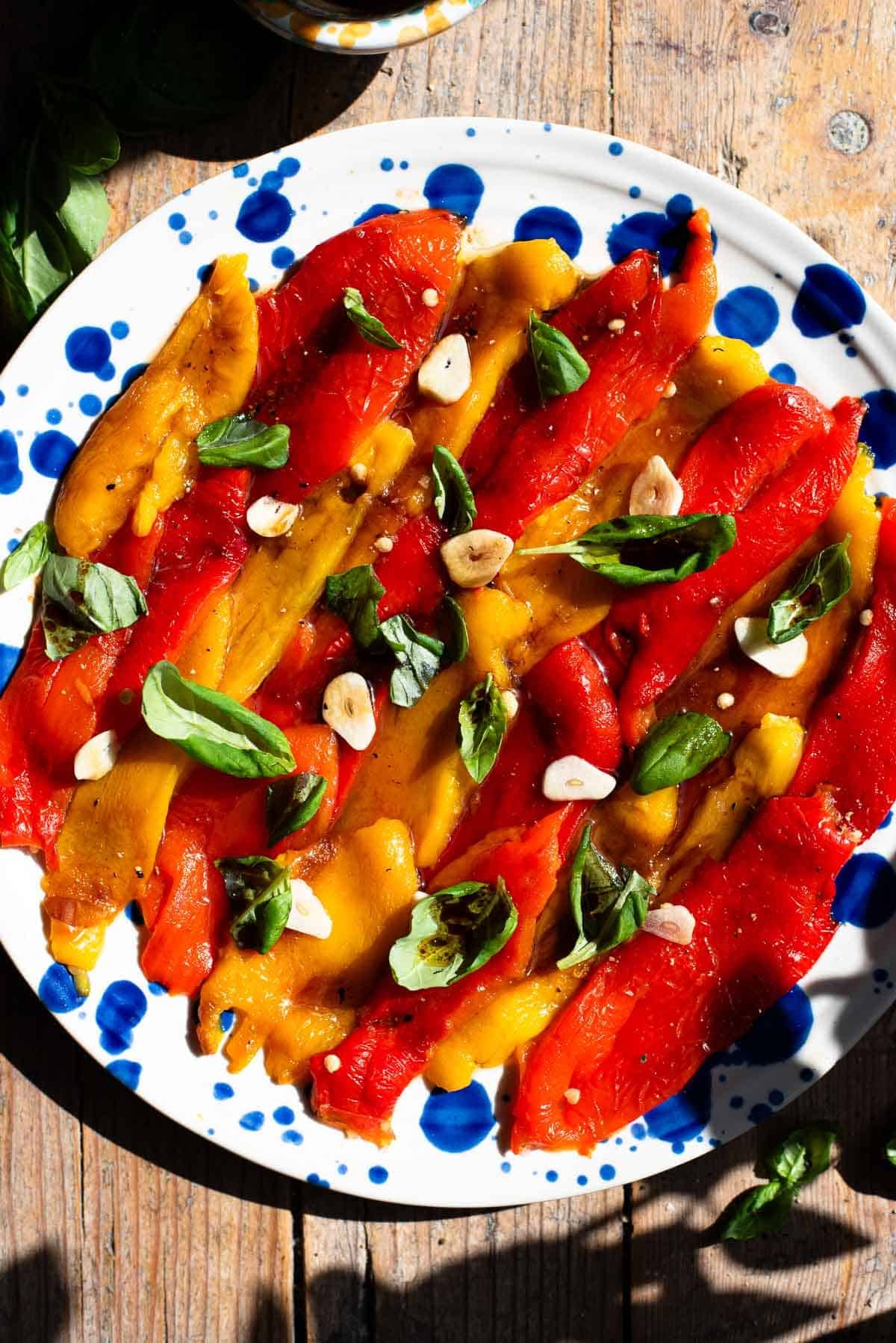 Red and yellow marinated roasted peppers arranged on a blue speckled plate topped with garlic and basil.