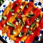 An overhead shot of marinated red and yellow bell peppers on a plate with garlic and basil. The background is rustic wood.
