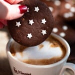 A close up of a hand holding one pan di stelle chocolate cookie over a cappuccino.