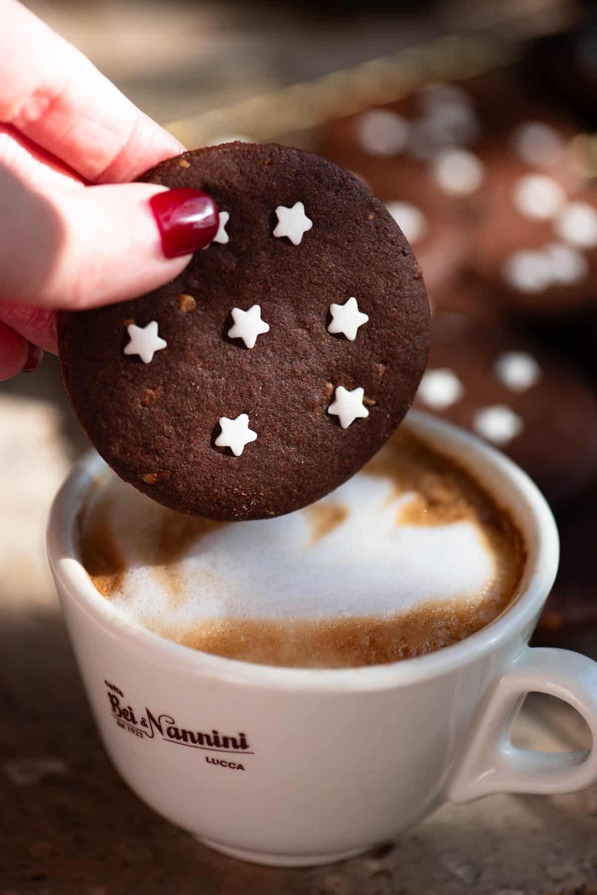 A close up of a hand holding one pan di stelle chocolate cookie over a cappuccino.