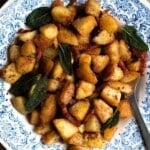 A close up cropped square image of crispy potatoes in a blue bowl with pancetta and sage.