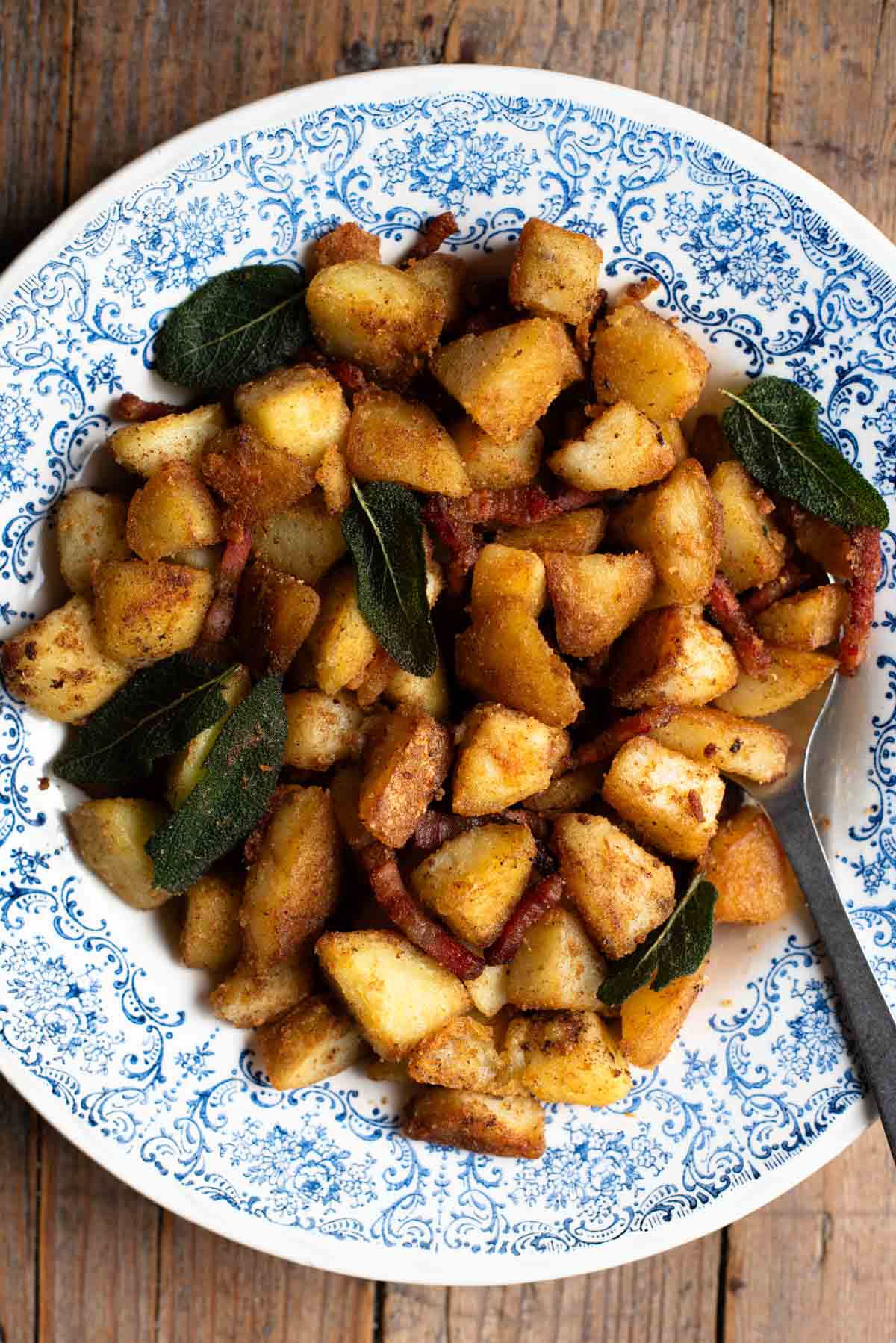 Crispy pan-fried potatoes with pancetta and sage in a blue and white patterned bowl on a rustic wooden surface.
