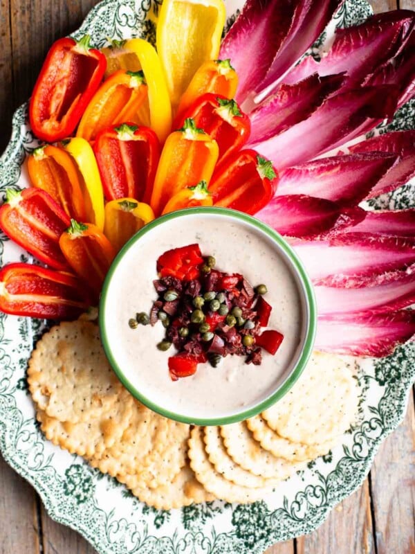 An overhead shot of a tuna dip topped with capers, pepper and olives on a platter with fresh peppers and chicory.