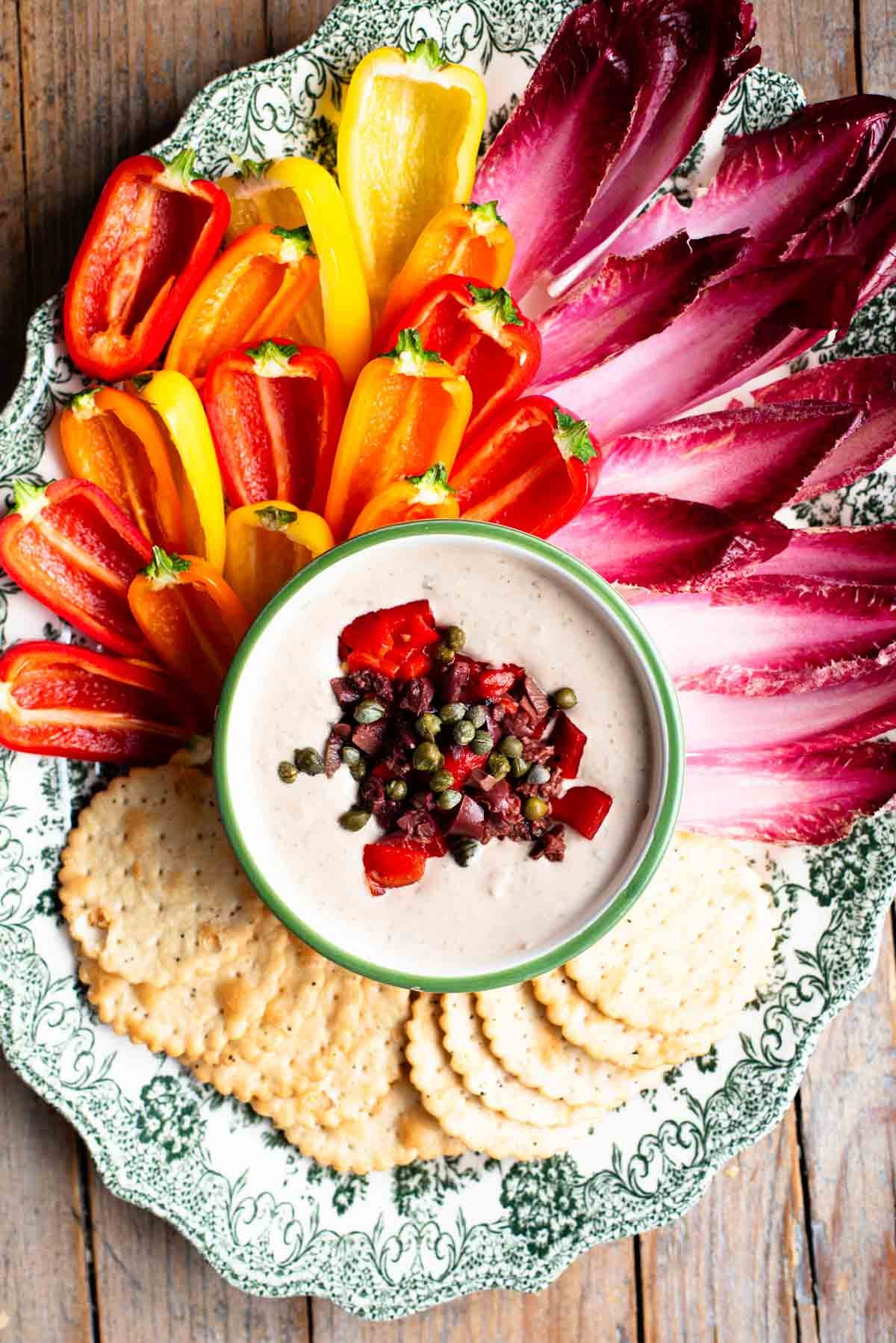 An overhead shot of a tuna dip topped with capers, pepper and olives on a platter with fresh peppers and chicory.