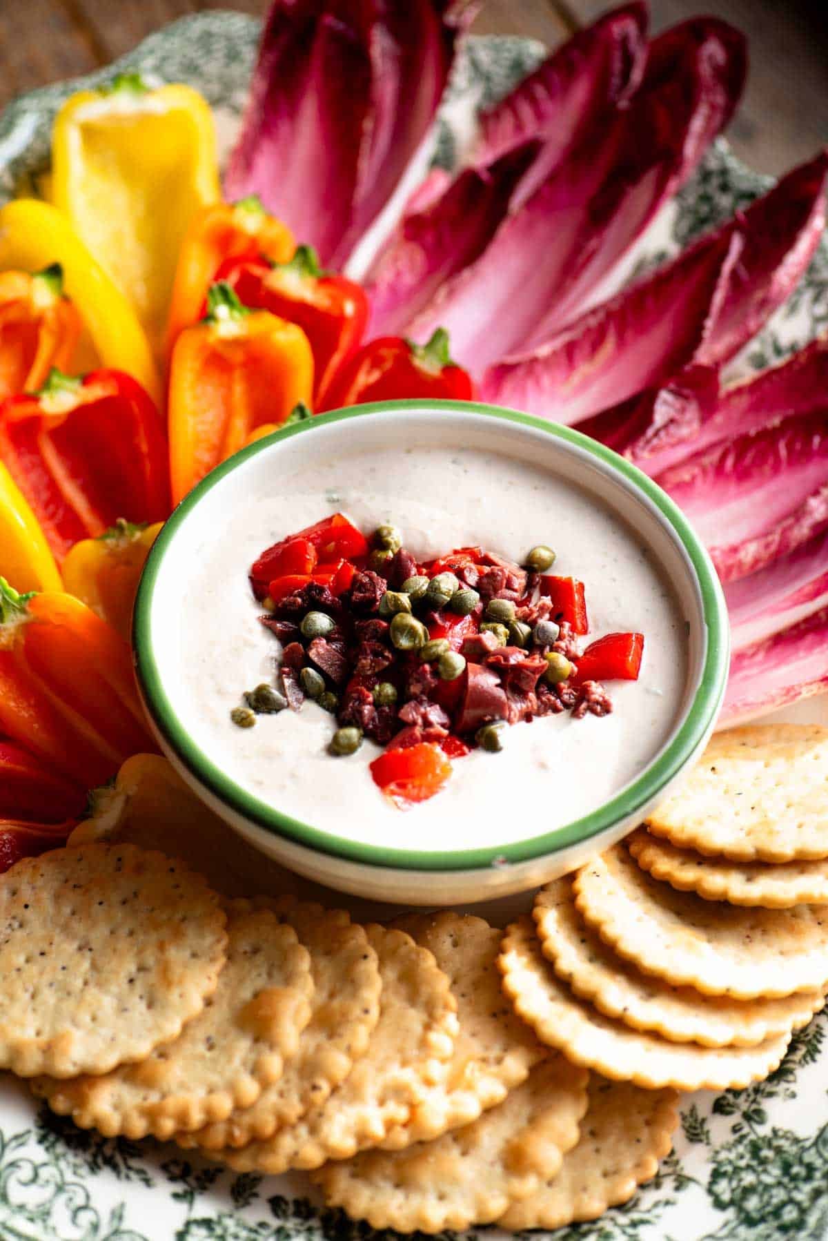 A close up side shot of a tuna dip topped with peppers, olives and capers on a colourful platter.